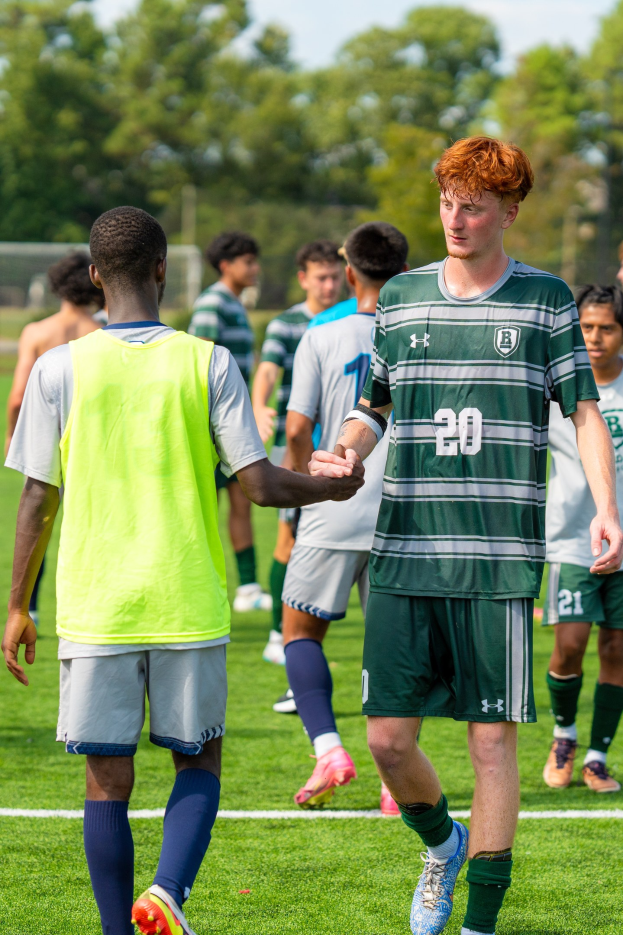 Junge Männer in Turnschuhen auf einem Fußballfeld unter einem klaren blauen Himmel, umgeben von Bäumen.