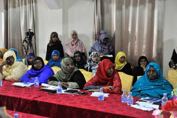 Gruppe von Frauen, die an einem Tisch mit verschiedenen Gegenst├Ąnden sitzen und diskutieren, mit einer Blumenvase rechts und Vorh├Ąngen im Hintergrund.