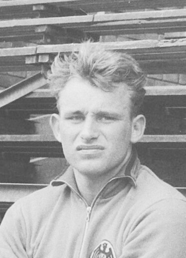 A black and white photo of an older man in a jacket standing in front of a pile of wooden planks.