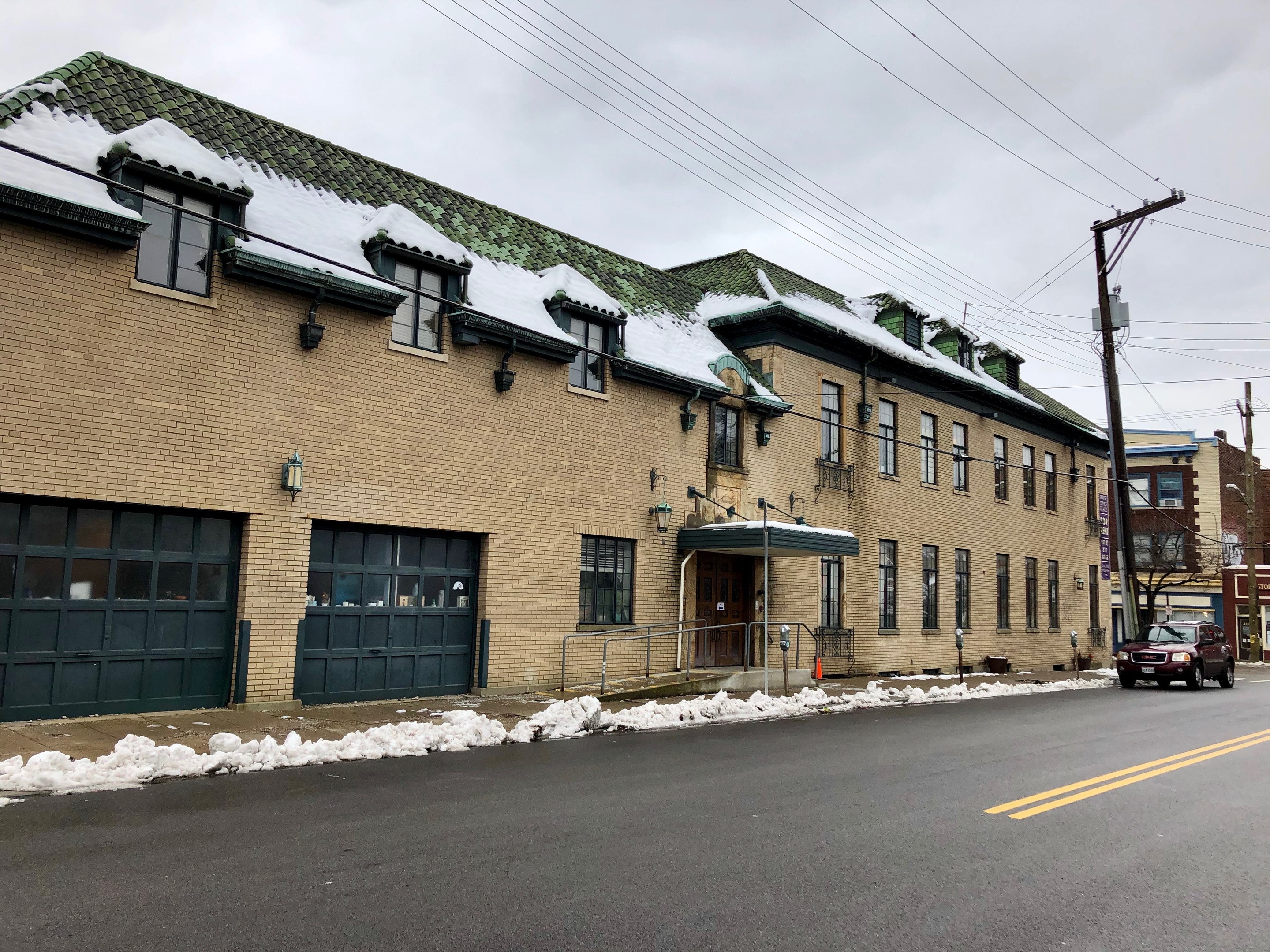 Großes Ziegelgebäude mit grüner Dachfläche an einer Straßenkreuzung, als ehemalige Feuerwache St. Louis bezeichnet, umgeben von Fahrzeugen, Schnee, Strommasten, Bäumen und einem bewölkten Himmel.