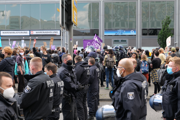 Große Menschenmenge protestiert vor einem Gebäude, einige halten Schilder und tragen Helme, mit einem Schildständer und einem Baum im Hintergrund.