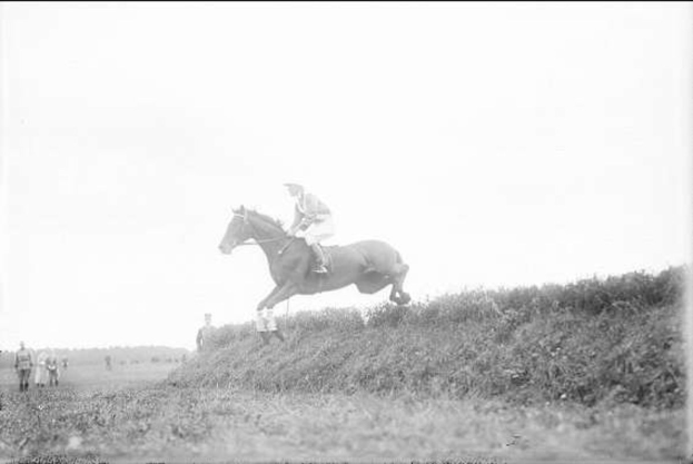 Schwarz-weißes Foto eines Pferdes und Reiters, die über einen grünen Hügel springen, mit wehender Mähne und Schweif des Pferdes und dem Reiter in Helm und langem Mantel, während Zuschauer im Hintergrund beobachten.