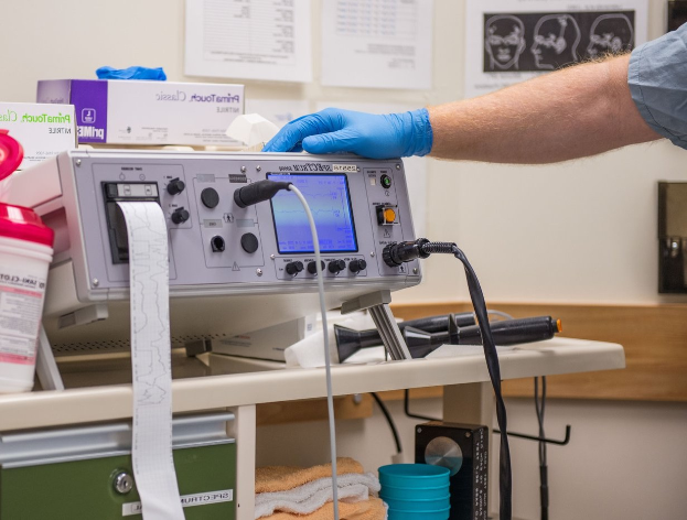 Eine Person in blauen Handschuhen arbeitet an einem medizinischen Gerät auf einem Tisch, umgeben von verschiedenen Gegenständen, mit Papieren an der Wand im Hintergrund, was auf eine Krankenhausatmosphäre hindeutet.