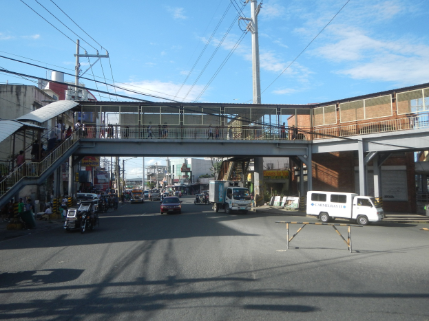 Stadtstraße mit Fahrzeugen, eine Fußgängerbrücke mit Menschen, Strommasten mit Drähten, Gebäuden und einem bewölkten Himmel.