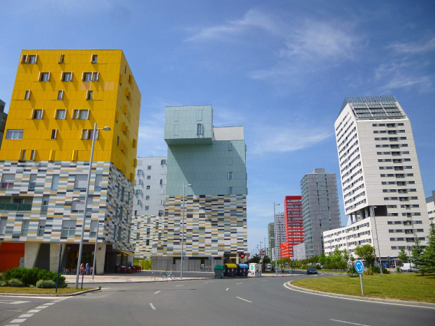 Eine Stadtstraße mit hohen Gebäuden im Hintergrund, Straßenlaternen, Straßenschildern, Autos, Informationsanzeigen, Büschen, Bäumen und einem bewölkten Himmel, mit den einzigartig gestalteten Gebäuden der Universität Rotterdam.