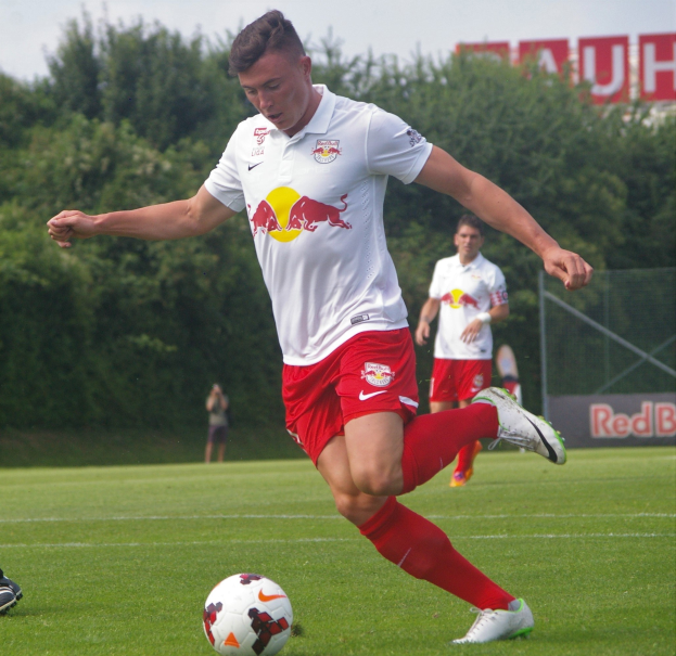 Berufsfußballer, der einen Fußball auf einem grünen Feld mit Bäumen und einem klaren blauen Himmel im Hintergrund kickt, Logo von RB Leipzig sichtbar.