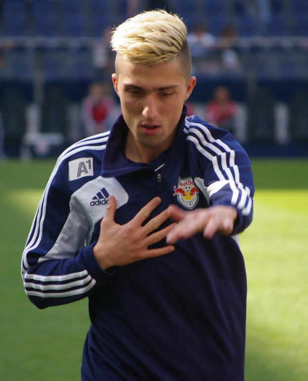 Ein junger Mann in einem blauen T-Shirt steht auf einem Fußballfeld und streckt die Arme aus, wobei er entschlossen wie ein Flügelspieler wirkt.