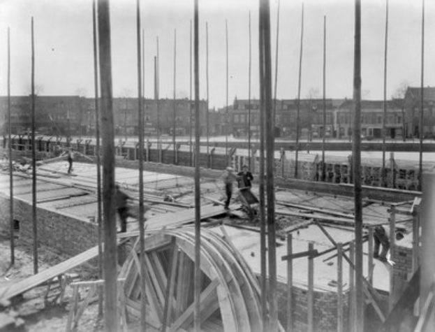 Schwarzes und weißes Foto einer Baustelle mit Arbeitern, hölzernen Pfählen, einem Rad im Vordergrund und Gebäuden, Bäumen und einem klaren Himmel im Hintergrund.