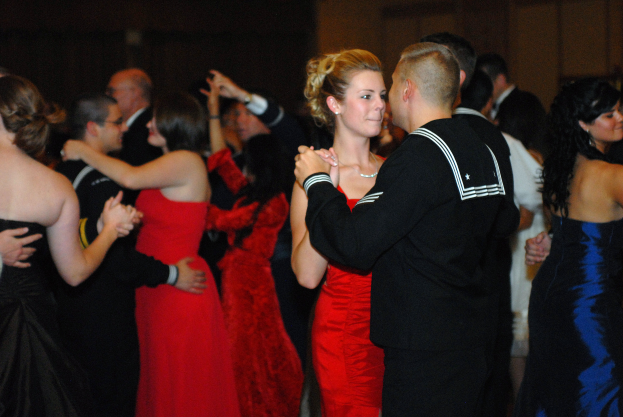 Eine Gruppe von Menschen in eleganten Kleidern tanzt energisch auf einer Tanzfläche mit einer Wand im Hintergrund.