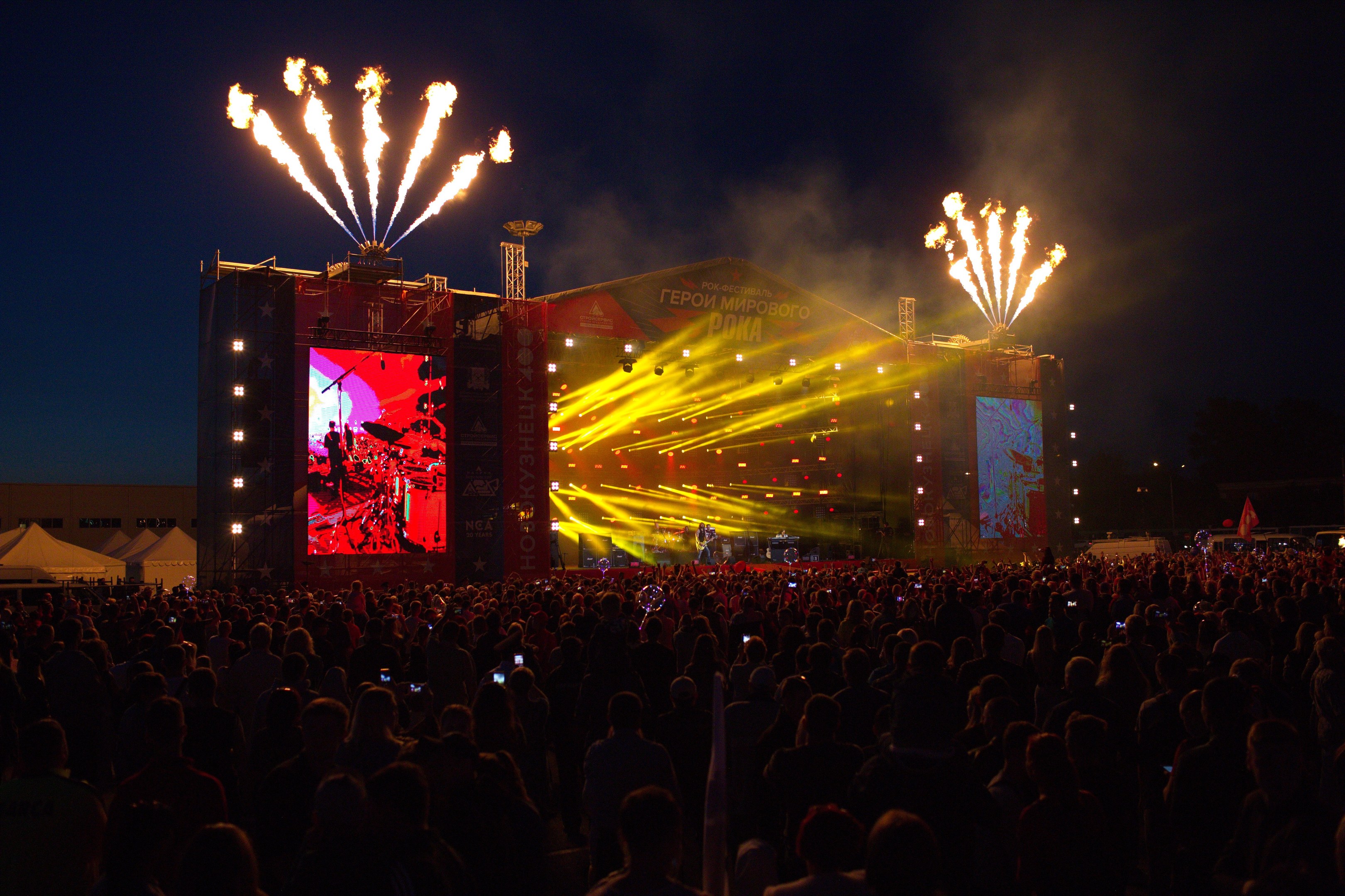 Große Menschenmenge auf einem Musikfestival, das eine bunt beleuchtete Bühne mit Zelten links daneben und einem Feuerwerk am dunklen Himmel zeigt.