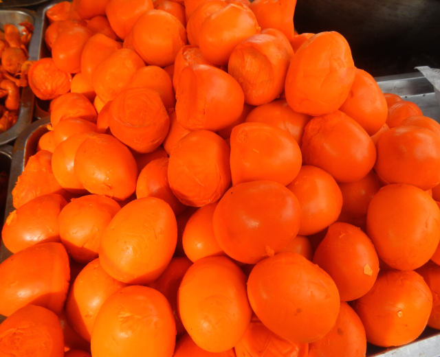 Ein Marktstand zeigt ein Metalltablett mit reifen, orangen Persimonen, daneben andere Lebensmittel und ein schwarzes Objekt in der oberen rechten Ecke.