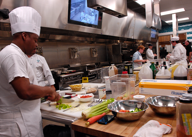 Eine Gruppe von Köchen in einer Küche bei der Zubereitung von Speisen auf einem Schneidbrett, mit einem Koch im Vordergrund in einer weißen Schürze und Mütze, der ein Messer hält und von verschiedenen Utensilien, Schüsseln, Gemüse, einem Handy, Flaschen und anderen Gegenständen umgeben ist, mit einem Fernseher und Deckenleuchten im Hintergrund.