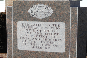 Gedenkstein mit einer Plakette für Feuerwehrleute in Millbury, vor einem Backsteingebäude mit umgebender Vegetation gesetzt.