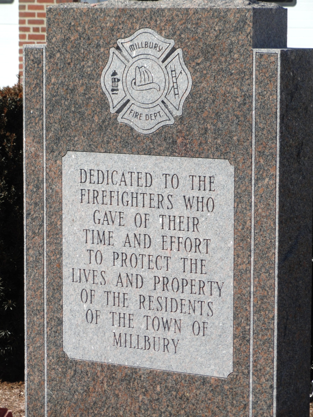 Gedenkstein mit einer Plakette für Feuerwehrleute in Millbury, vor einem Backsteingebäude mit umgebender Vegetation gesetzt.