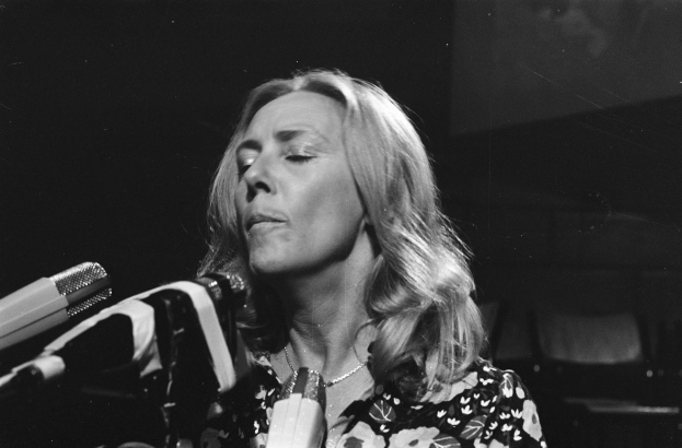 Eine Frau, die leidenschaftlich in ein Mikrofon singt, trägt ein schwarzes und weißes Outfit mit einem klassischen Bob-Haarschnitt, vor einem dunklen Hintergrund.