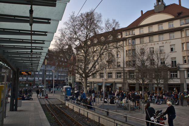 Lebendige Stadtstraße mit einem Bahnhof auf der linken Seite, umgeben von Gebäuden, Bäumen und Laternen, mit Menschen, die gehen, sitzen und stehen, in der Nähe des Bahnhofs unter einem sichtbaren Himmel.
