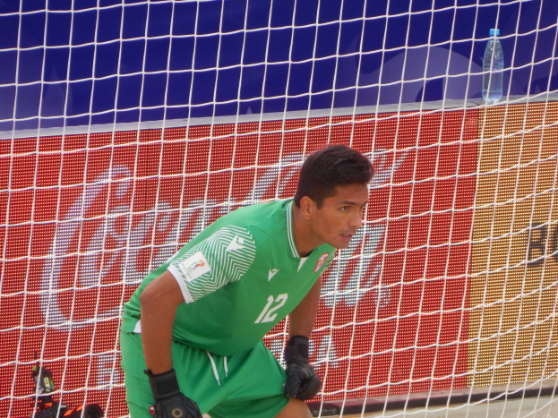 Ein Torwart in grüner Uniform kniet vor einem Tor mit Netz im Hintergrund und einer Flasche rechts daneben.