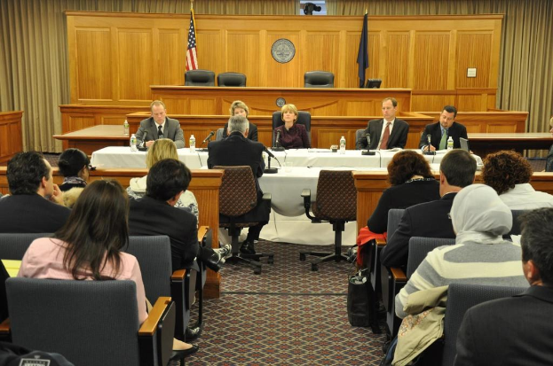 Versammlungssaal mit einer Gruppe von Menschen, die auf Stühlen sitzen, zwei ausländische Flaggen, ein orangefarbener Schreibtisch mit Wasserflaschen und ein Mikrofon.