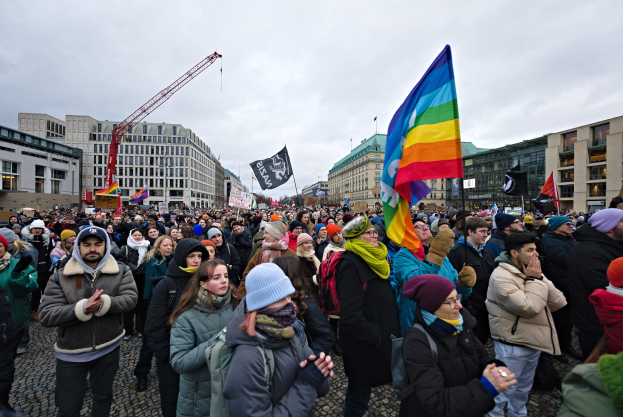 Große Gruppe von Menschen bei einer LGBTQ+-Rechtsdemo in Berlin, die Fahnen und Schilder mit Text halten, vor einem Gebäude mit Fenstern, einem Kran und einem bewölkten Himmel.