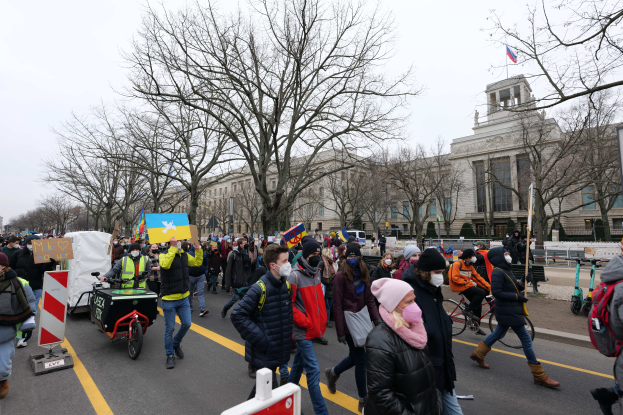 Eine große Gruppe von Menschen nimmt an einer Protestmarsch auf einer Straße in Washington, D.C. teil, einige halten Plakate und Banner, andere fahren Fahrräder und es gibt Schilder, Bäume und einen klaren blauen Himmel im Hintergrund.