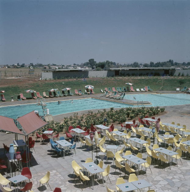 Ein Freibad mit Tischen, Stühlen und Sonnenschirmen, umgeben von Blumenpflanzen, einem Zaun, einer Wand, Bäumen und Gebäuden unter einem bewölkten Himmel.