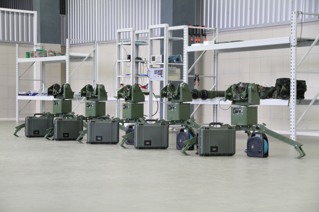 Eine Gruppe von Militärrobotern ist auf einem Boden umgeben von Koffern und anderen Gegenständen auf Gestellen positioniert, mit einer Wand und einem Geländer im Hintergrund.