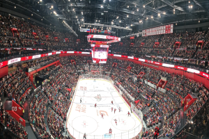 Ein großes, hell erleuchtetes Stadion voller jubelnder Zuschauer, die ein Hockey-Spiel sehen, mit einem Bildschirm in der Mitte der Eisfläche.