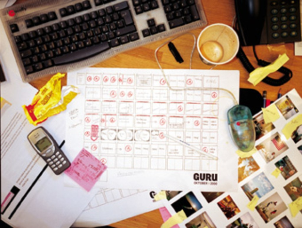 Ein chaotischer Schreibtisch mit Papieren, einem Computer, einem Telefon, Tassen und einem Handy.