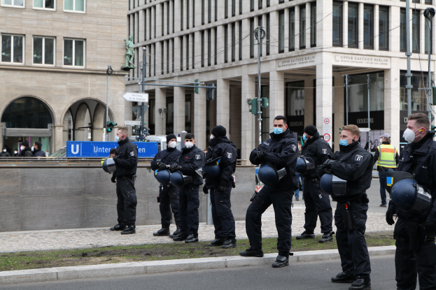 Eine Gruppe von Polizisten in schwarzen Uniformen und Masken steht vor einem Glasfenstergebäude mit Säulen in Berlin, Deutschland, einige halten Helme, mit Laternen, Verkehrsampeln, Texttafeln, einer Statue, anderen Menschen und einem grasbewachsenen Boden im Hintergrund.