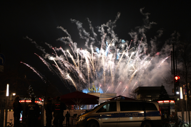 Ein Polizeiwagen vor einer Menschenmenge während einer Silvesterfeier in Berlin, mit farbigen Feuerwerken am Himmel und Stadtlichtern.