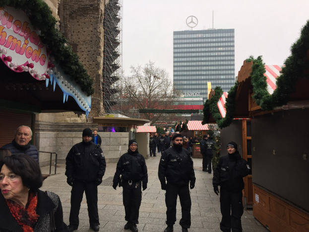 Polizeibeamte in schwarzen Uniformen stehen vor einem geschmückten Weihnachtsmarkt in Berlin, mit festlichen Dekoelementen, Gebäuden, Bäumen und einem klaren blauen Himmel im Hintergrund.