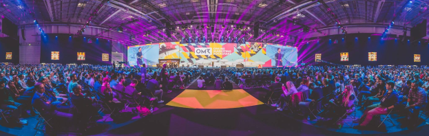 Eine große Zuschauermenge sitzt auf Stühlen einem hell erleuchteten Bühne mit mehreren Lautsprechern und Bildschirmen gegenüber, wahrscheinlich bei einem Online-Marketing-Event.