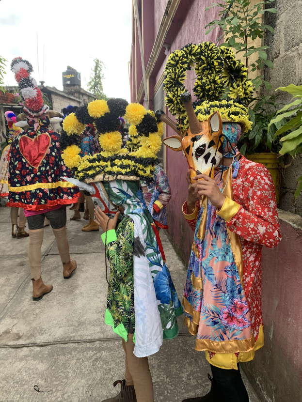 Eine Gruppe von Menschen in bunten Kostümen steht vor einem Gebäude während eines Faschingsumzugs, haltend verschiedene Gegenstände mit Bäumen und Gebäuden im Hintergrund unter einem klaren blauen Himmel.