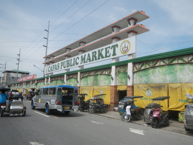 Eine belebte Stadtstraße mit Autos, Motorrädern und Rikschas, die an dem Gebäude "Capas Public Market" vorbeifahren, vor dem Hintergrund von Strommasten, Laternenmasten, Gebäuden und einem bewölkten Himmel.