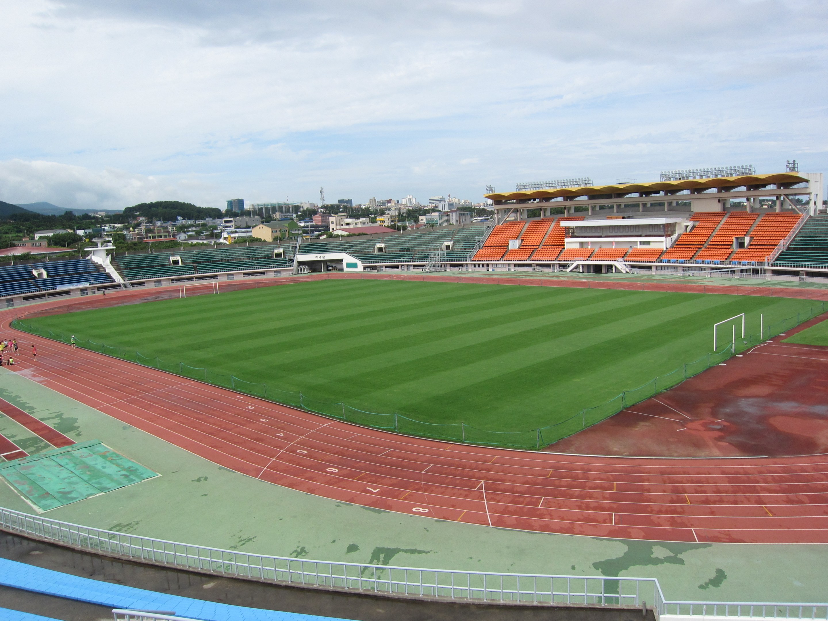 Großes Stadion mit einem Fußballfeld, umgeben von Gebäuden, Bäumen, Hügeln und einem klaren blauen Himmel, mit einigen Menschen und saftigem Grün.