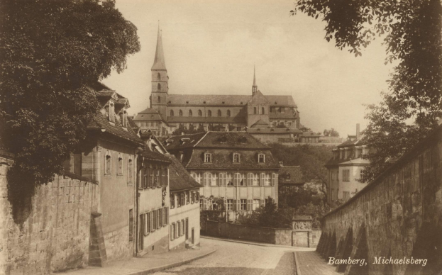 Schwarz-weißes Foto einer Stra├če in Bamberg, Deutschland, mit einer Kirche im Hintergrund, flankiert von Mauern und gesäumt von B├Ąumen.