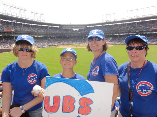Drei Frauen in Kappen und Brillen, ein Junge in Brille und Kappe mit einem Ball und einer Tafel, stehend in einem Stadion mit Publikum und Feld dahinter, unter einem Himmel mit wenigen Lichtern.