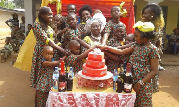 Eine Gruppe von Menschen, die um einen Tisch mit einem Kuchen in einer ländlichen Outdoor-Umgebung feiern, mit Häusern, Bäumen und einem klaren blauen Himmel im Hintergrund.