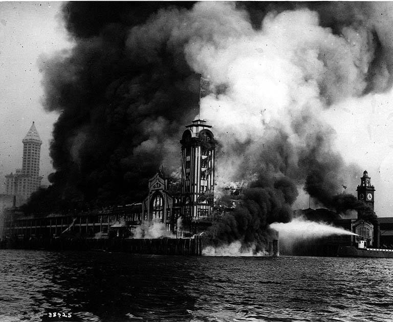 Schwarzes und weißes Foto eines großen brennenden Gebäudes mit Rauch, der herausquillt, einem Gewässer im Vordergrund und anderen Gebäuden und dem Himmel im Hintergrund.