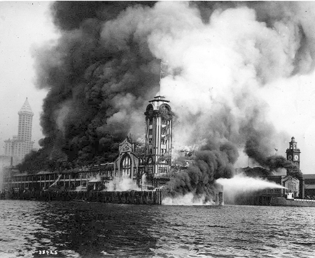 Schwarzes und weißes Foto eines großen brennenden Gebäudes mit Rauch, der herausquillt, einem Gewässer im Vordergrund und anderen Gebäuden und dem Himmel im Hintergrund.