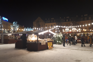 Ein nächtlicher Weihnachtsmarkt in einer verschneiten Straße mit Menschen, Kinderwagen, beleuchteten Hütten, Pflanzen, Bäumen, Gebäuden und Schildern unter einem bewölkten Himmel.