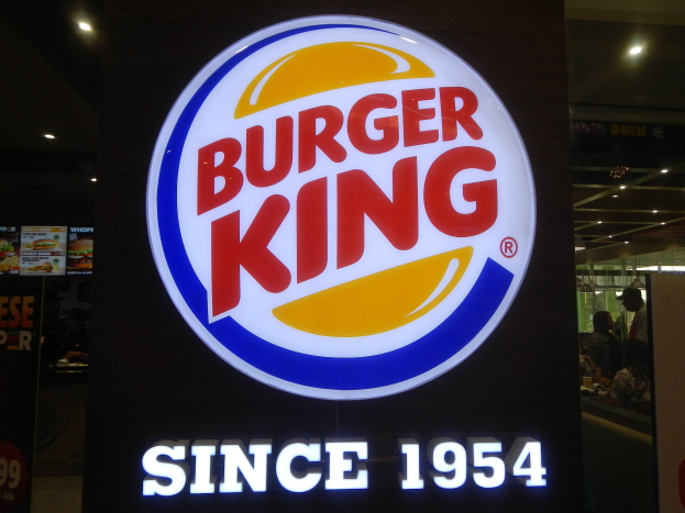 Burger King-Schilder in einem Restaurant mit Menschen und anderen Gegenständen sowie einer Tafel mit dem Burger King-Logo und Text links daneben.