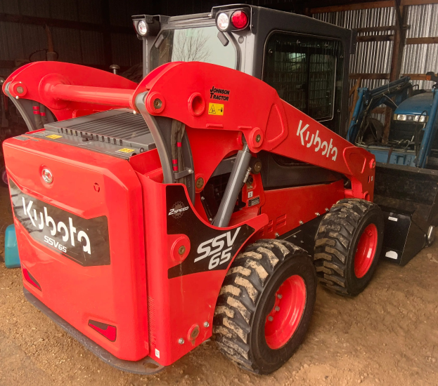 Ein roter Kubota SSV65 Radlader steht vor einer Hütte, mit anderen Fahrzeugen im Hintergrund.