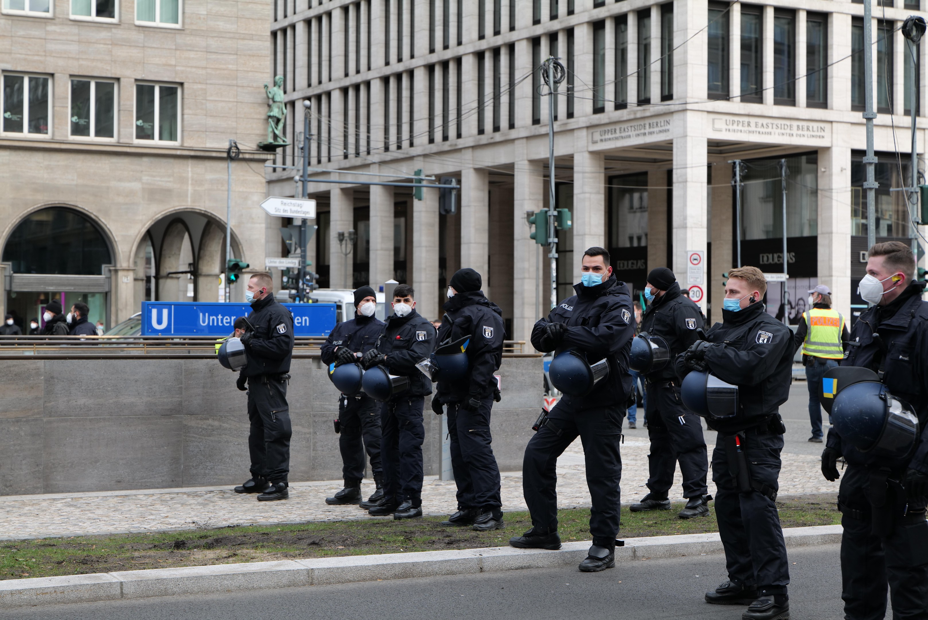 Eine Gruppe von Polizisten in schwarzen Uniformen und Masken steht vor einem Glasfenstergebäude mit Säulen in Berlin, Deutschland, einige halten Helme, mit Laternen, Verkehrsampeln, Texttafeln, einer Statue, anderen Menschen und einem grasbewachsenen Boden im Hintergrund.