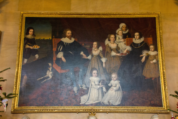 Gemälde einer Familie in einem Raum mit einem Weihnachtsbaum, gerahmt und an einer Wand mit zwei Fotorahmen daneben aufgehängt, und dekorative Gegenstände unten.