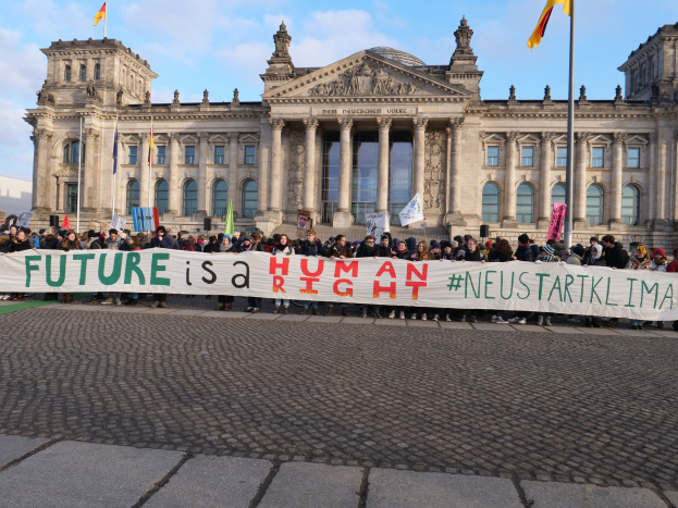 Eine Gruppe von Menschen hält ein Transparent mit der Aufschrift "Zukunft ist ein Mensch" vor dem Reichstaggebäude in Berlin, Deutschland, mit den architektonischen Details des Gebäudes und Flaggen im Hintergrund bei bewölktem Himmel.