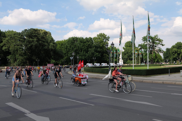 Gruppe von Radfahrern auf einer von Bäumen gesäumten Straße mit Laternenmasten und Flaggenmasten während der Pariser Radtour 2016, mit Fahrzeugen und bewölktem Himmel im Hintergrund.