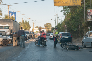 Eine Gruppe von Menschen umringt ein verunglücktes Motorrad am Straßenrand mit mehreren Fahrzeugen, darunter ein Lastwagen, im Hintergrund und Bäumen, Mästängen, Lichtern, Schildern und dem Himmel.