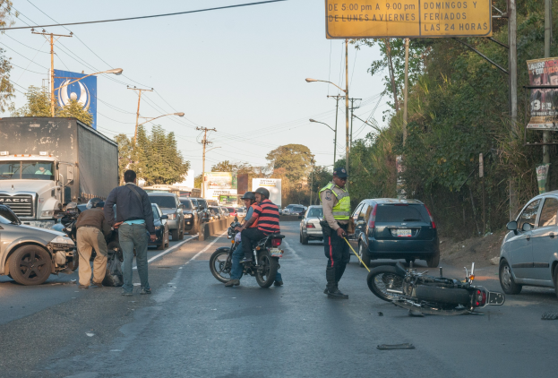 Eine Gruppe von Menschen umringt ein verunglücktes Motorrad am Straßenrand mit mehreren Fahrzeugen, darunter ein Lastwagen, im Hintergrund und Bäumen, Mästängen, Lichtern, Schildern und dem Himmel.