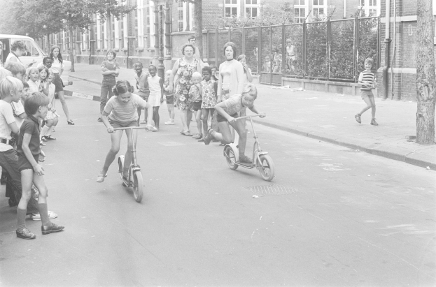 Schwarzes Bild von Kindern, die auf Roller die Straße hinunterfahren, mit einem geparkten Auto auf der linken Seite, Gebäuden, Bäumen, Pfählen und einem Zaun im Hintergrund.